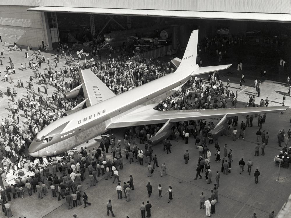 Top view of commercial jet with four engines and Boeing livery. People are crowded around the prototype jet.