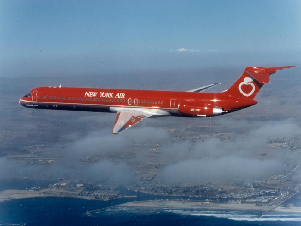 Side view of commercial jet in flight. Aircraft features red and white New York Air livery and two jet engines near the back of the plane.