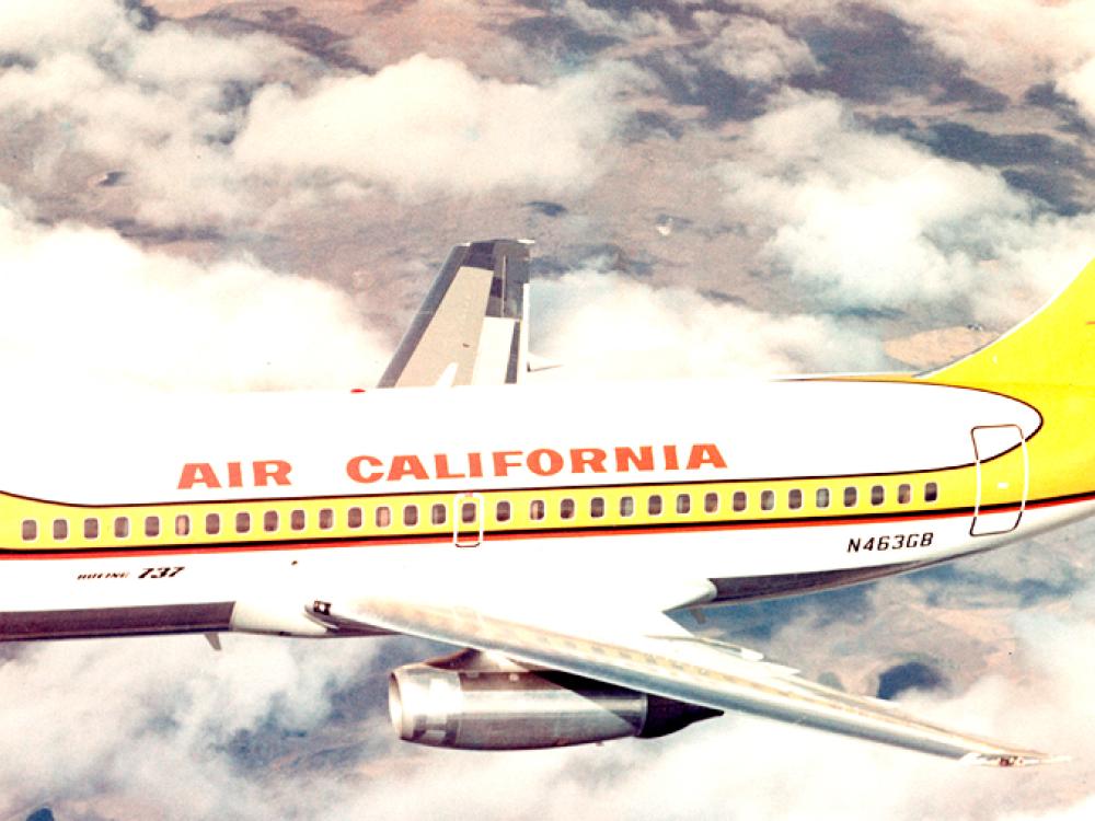 Side view of commercial aircraft in flight. Aircraft features two engines and white, yellow, black, and red Air California livery.