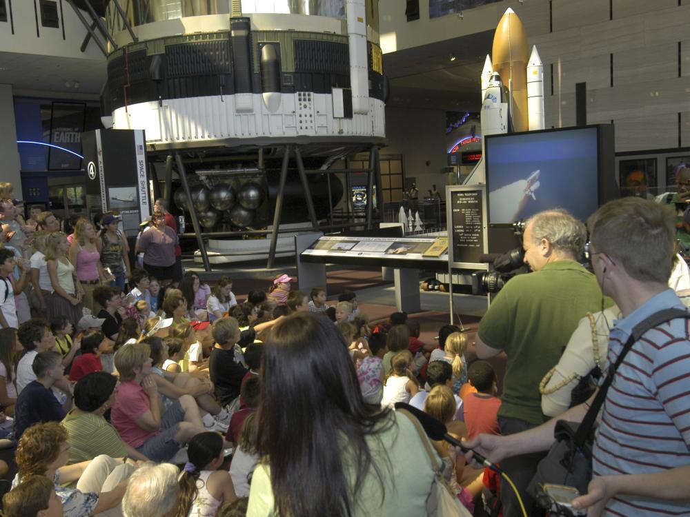 Museum visitors gathered around a large TV with Space Shuttle launch visible on the screen.