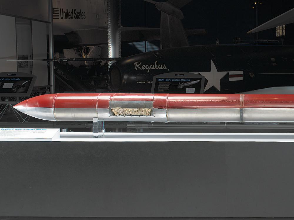 A long and skinny cylinder with a pointed tip at one end and tail fins at the other end. One half is painted red and the other half is left unpainted, showing the silver metal material it is constructed of.