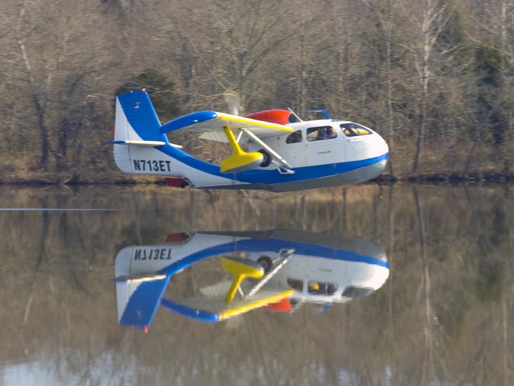 Side view of blue and white amphibious aircraft flying slightly above body of water. Registration number N713ET is painted in blue on the lower part of the vertical stabilizer.