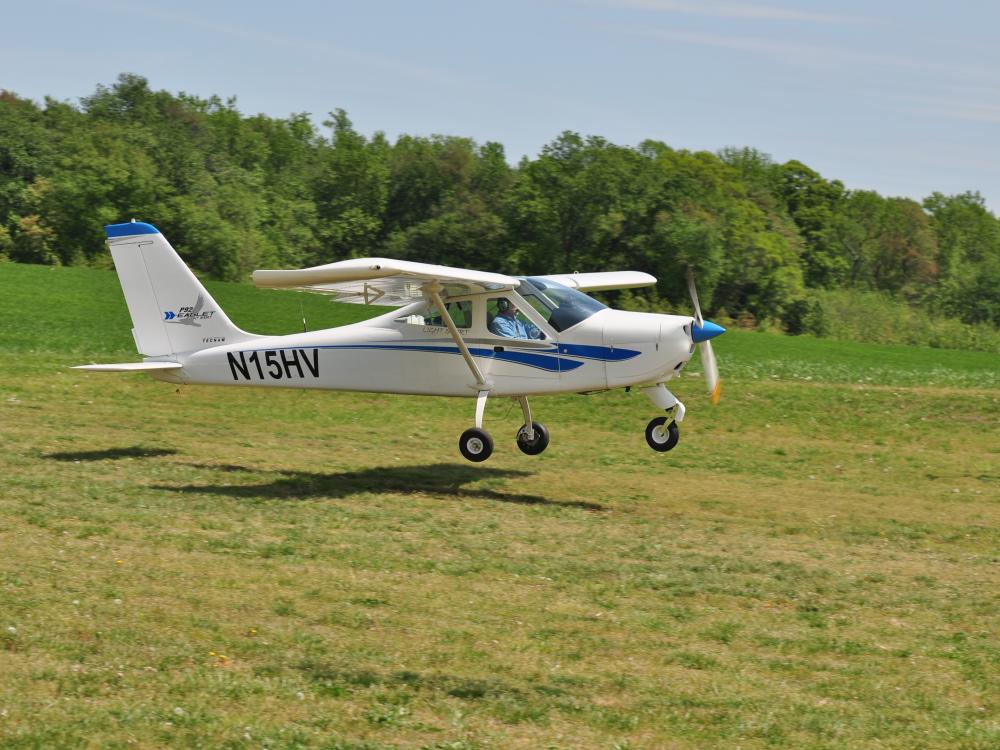 A white monoplane with a single engine flies just above the ground. The registration number "N15HV" is written in black near the rear of the fuselage.