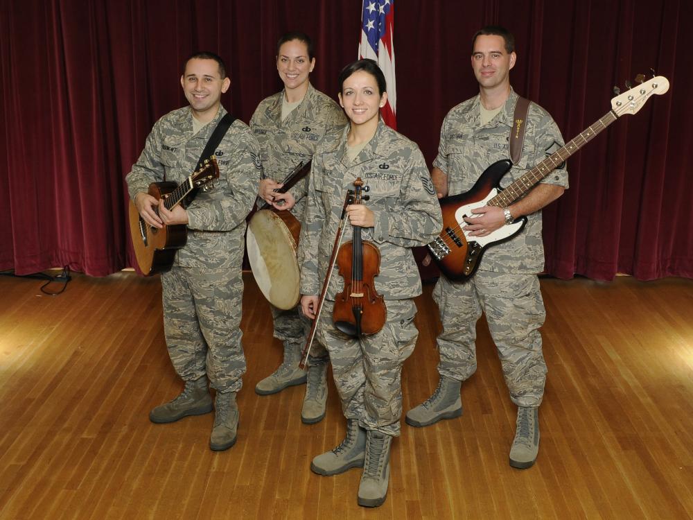 A group of four people pose together in U.S. Air Force camouflage uniforms. Each is holding an instrument: Two hold guitars, one plays a banjo, and the fourth plays the violin.