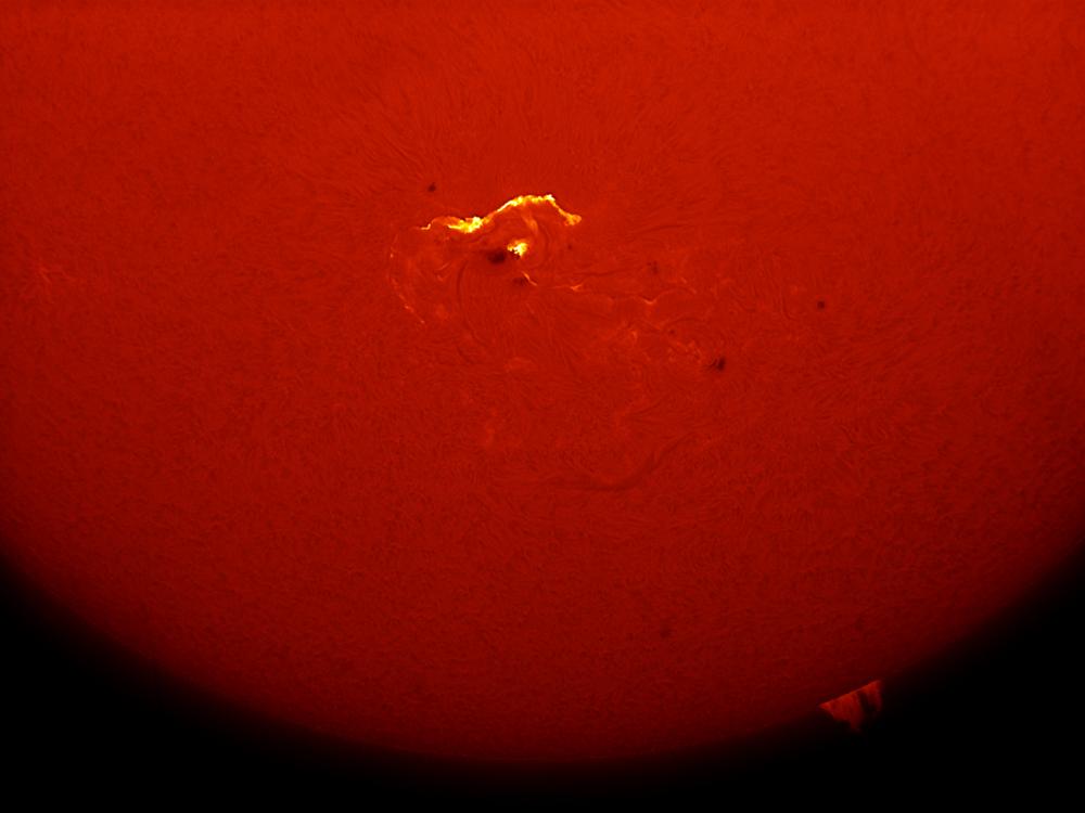 Partial disk view of the Sun focusing on a solar flare, highlighted in yellow with nearby black darkspots, visible from an image of the chromosphere of the Sun.