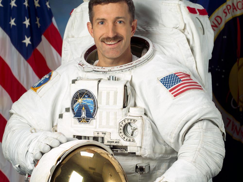 Dan Burbank, a male astronaut, stands for a photo wearing astronaut gear. He is standing in front of a background featuring a U.S. red, white, and blue flag and a second flag featuring NASA's seal.