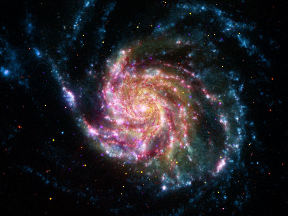 A still image of a spiral galaxy similar to the Milky Way galaxy, but larger in size. This galaxy features blue, white, pink, red, and yellow colors.