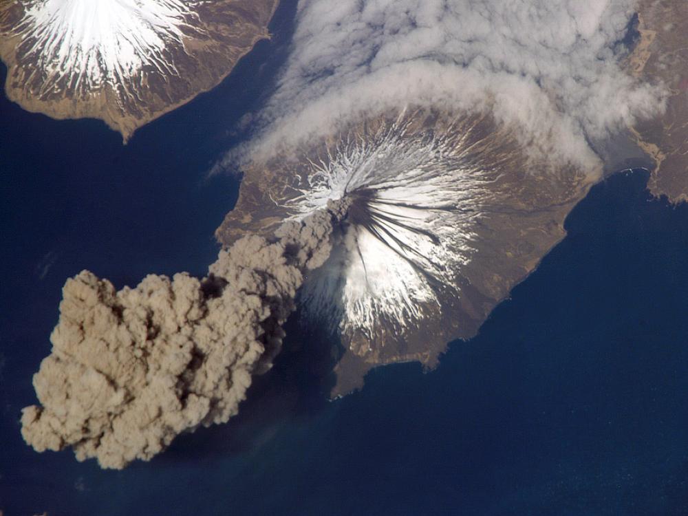 View from space of a volcanic eruption. A gray cloud of ash seen in the bottom left corner is floating away from the volcano site.