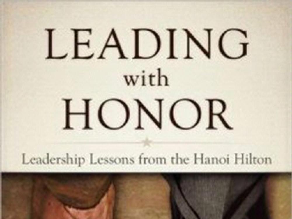 Cover of a book titled "Leading with Honor: Leadership Lessons from the Hanoi Hilton". Below the title is an image of two feet. The right foot is bare without any shoes whereas the left foot is wearing a black dress shoe.