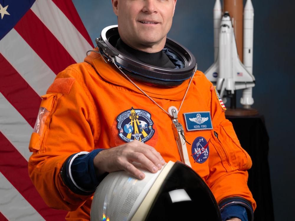 Commander Kevin Ford, a white male astronaut, poses for his official astronaut portrait in an orange suit. He is holding an astronaut helmet.