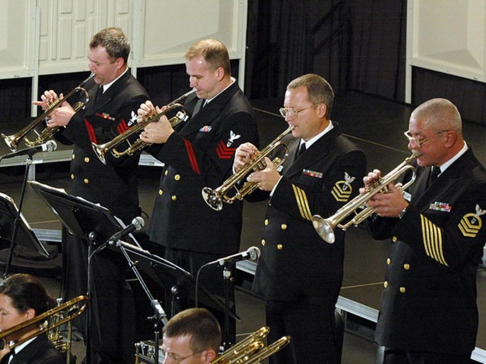 A group of musicians in U.S. Navy uniforms perform using various brass instruments.