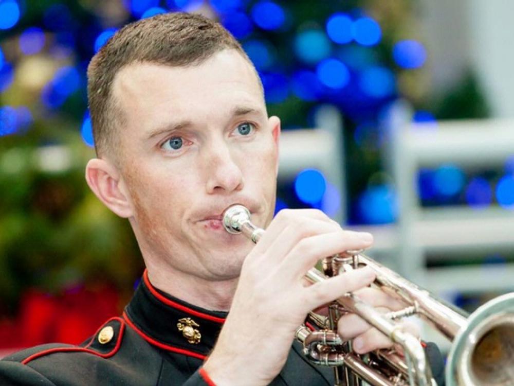 A man plays a trumpet in Marine Corps uniform as part of a larger group.