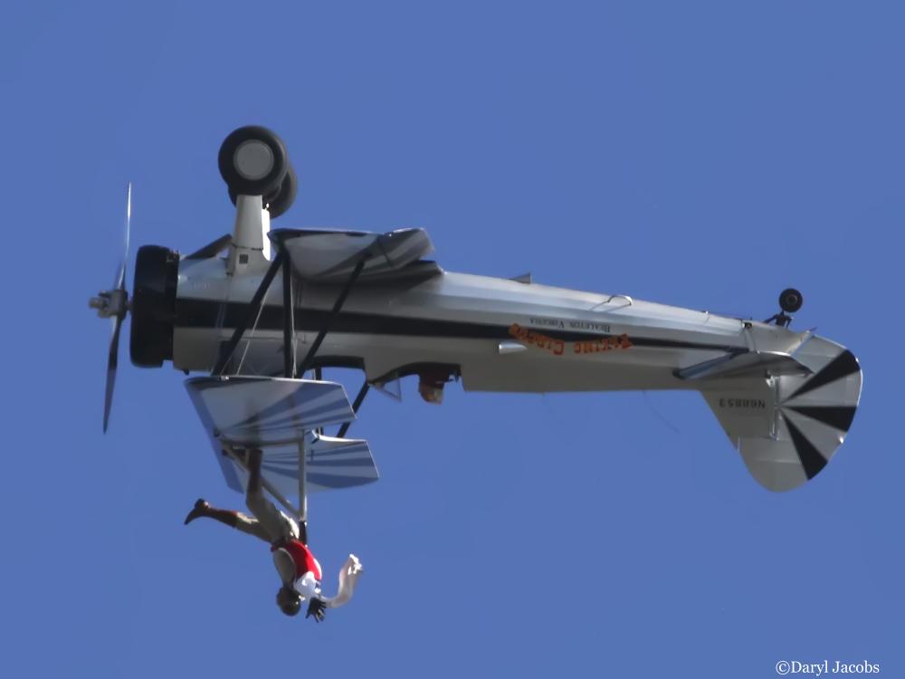 Side view of white and black biplane with single engine flying upside down. A person is depicted as standing on the upper wing of the plane.