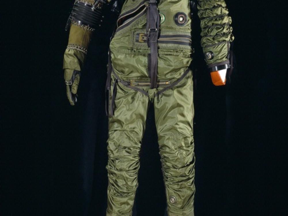 Front view of full-body spacesuit made of dark green-colored material. An opening for the helmet is present and reveals an orange-colored bladder. A black material covers the right shoulder and forearm.