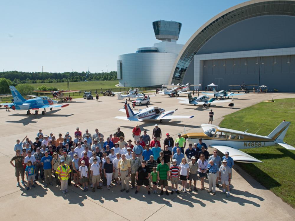 A group of pilots and museum staff pose as a group on a runway with planes behind them.