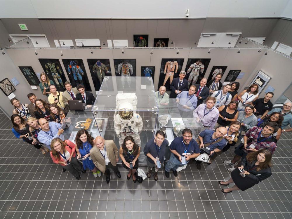 A large group of people stand around an museum exhibit featuring spacesuits.