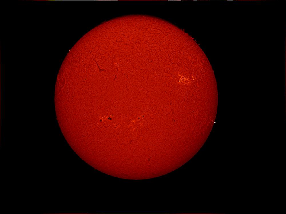 Disk view of the Sun's atmosphere with sunspot clusters (dark spots) surrounded by phages of lighter-colored clouds. A dark line known as a filament is also visible on the top left quadrant of the disk.