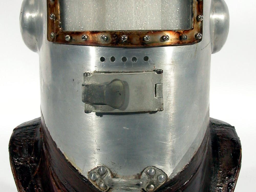 A primitive pressure helmet which looks like a knight's helmet with metal full-body face guard and darker-colored brim for attachment to the rest of a pressurized suit.