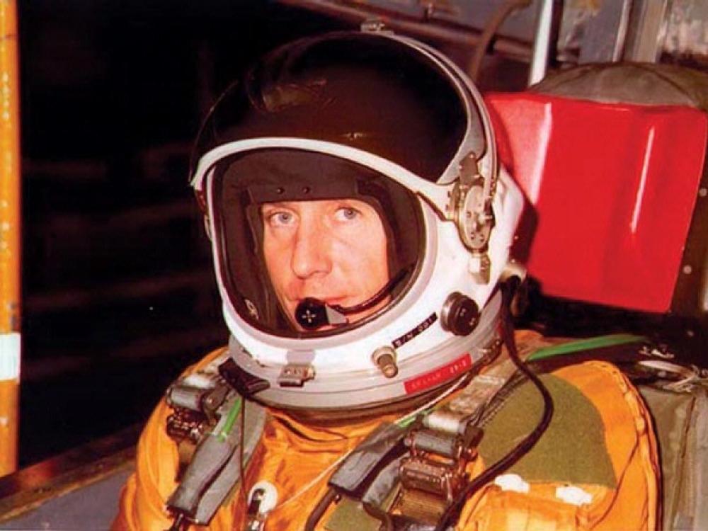 Col. Richard Graham, a white man, sits in an orange and green flight suit and white helmet.