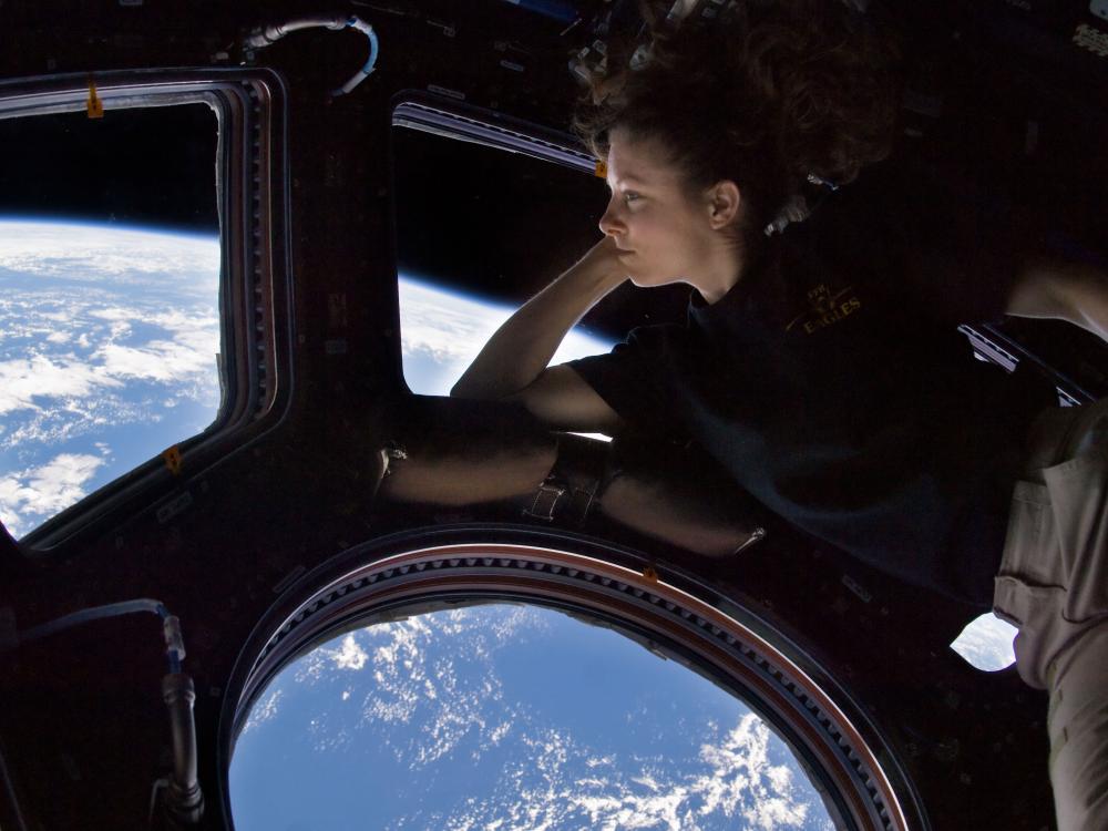 Tracy Caldwell Dyson, a white female astronaut, leans on a window showing the Earth in space.