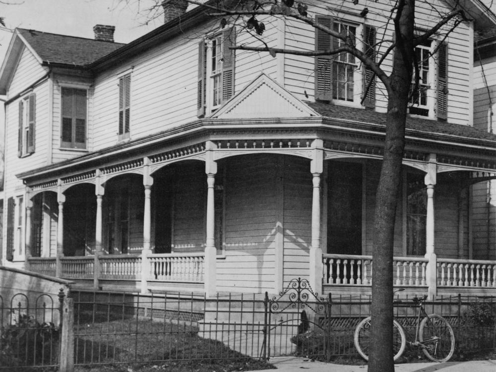 A large white house with a wrap around porch. A bicycle is leaned up against a short iron fence which surrounds the yard.