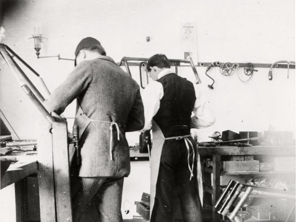 Two men with their back turned to the camera, working at a work bench and surrounded by tools.