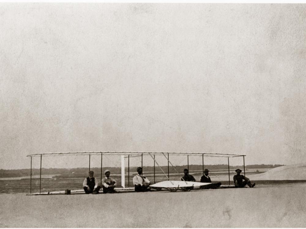 Six men sit on the ground in front of a glider