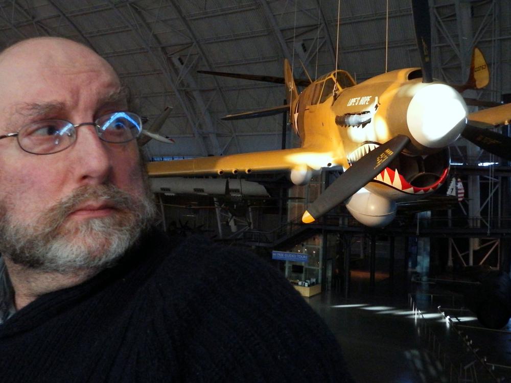 A white male specialist at the museum poses in front of a yellow monoplane with a shark mouth painted on the lower nose.