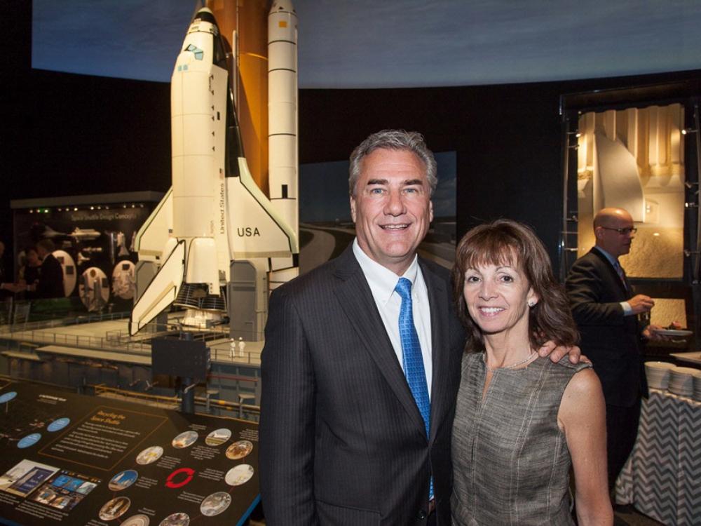 Two people, a white male and a white woman, pose together with a backdrop of a space shuttle model behind them.