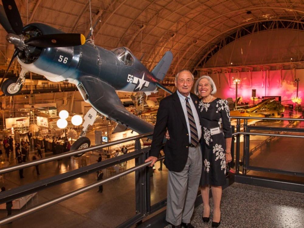 Robert and Lenore Briskman, a couple consisting of a white man and woman, pose together with the backdrop of a gray and white monoplane behind them.