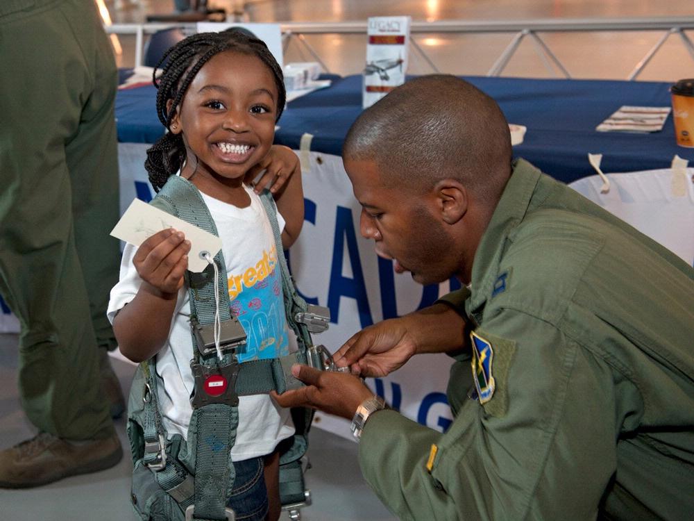 A young girl smiles as she receives assistance from a man with trying on a parachute at a museum event.