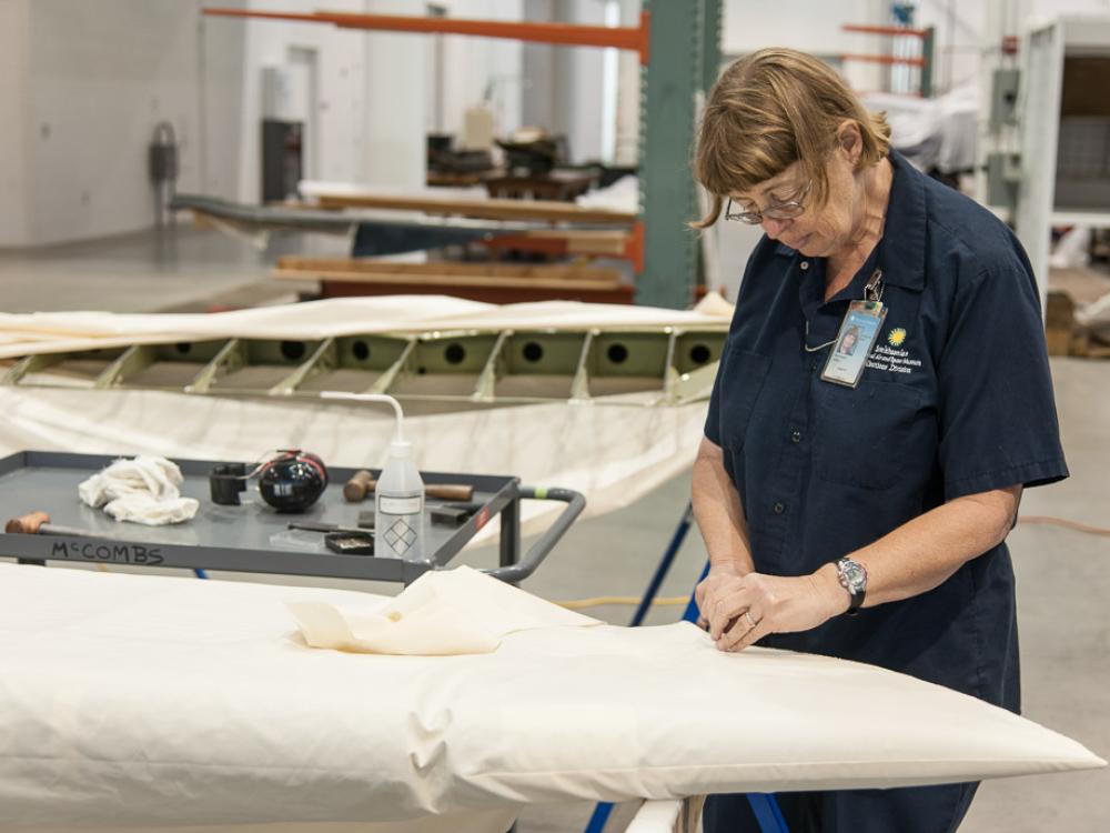 A white female museum specialist performs restoration work involving fabric on the Curtiss Helldiver, an aircraft.