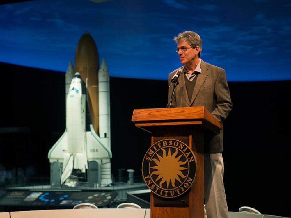 A white man who is a Curator of Space History at the Museum speaks at a podium. A backdrop includes a space shuttle.