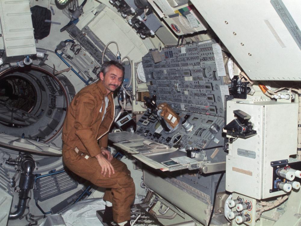 Owen Garriott, a male astronaut, stands near a console inside a space station.