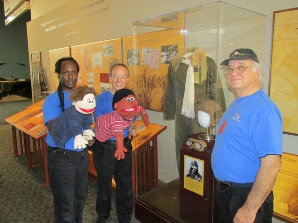 Three people, two of which are holding puppets, stand together in front of a museum display.