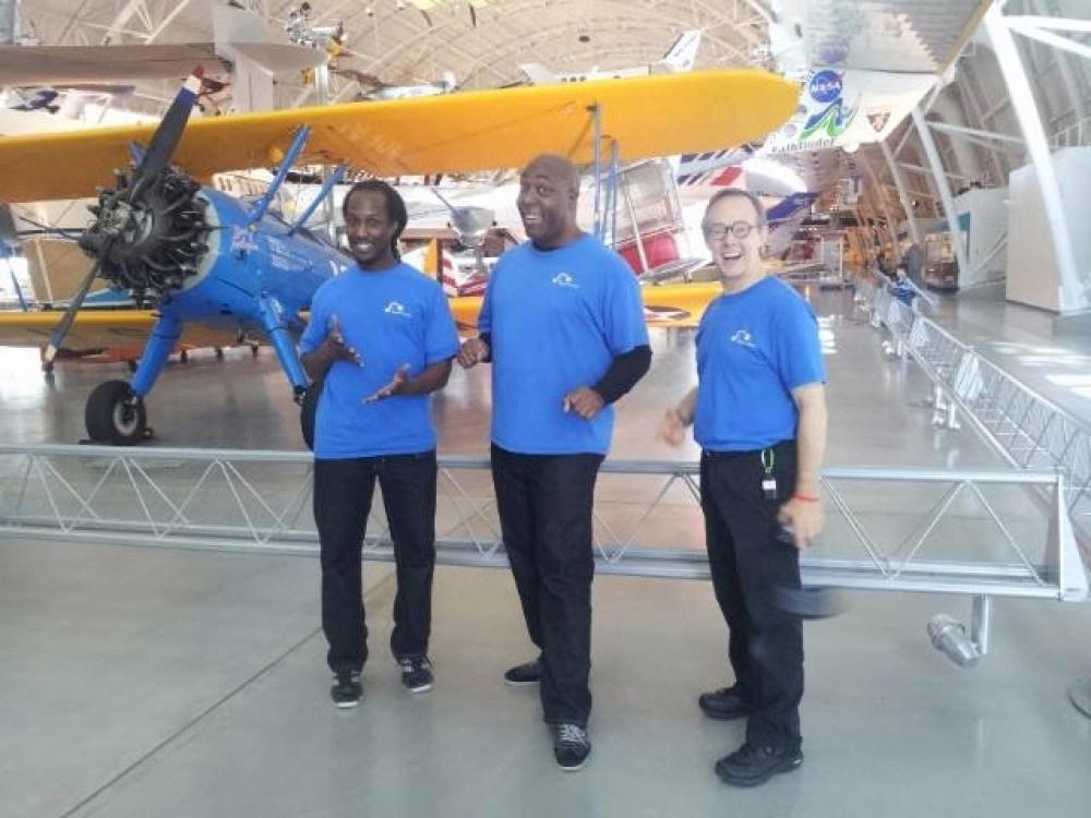 Three people who work as part of a puppetiering theater company stand together in front of a yellow and blue biplane inside the Museum's Steven F. Udvar-Hazy Center.