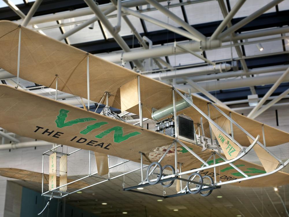 Biplane made with tan fabric and white metal hanging in the museum. "Vin Fiz" written on the bottom part of the lower wing in green paint.