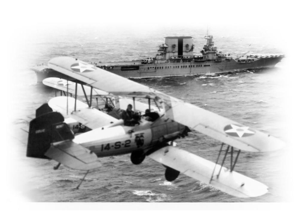 A military biplane flies over the ocean near an aircraft carrier.