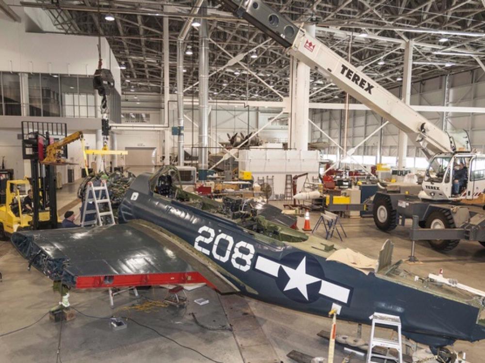 The engine of a blue military monoplane is placed by crane into the aircraft.