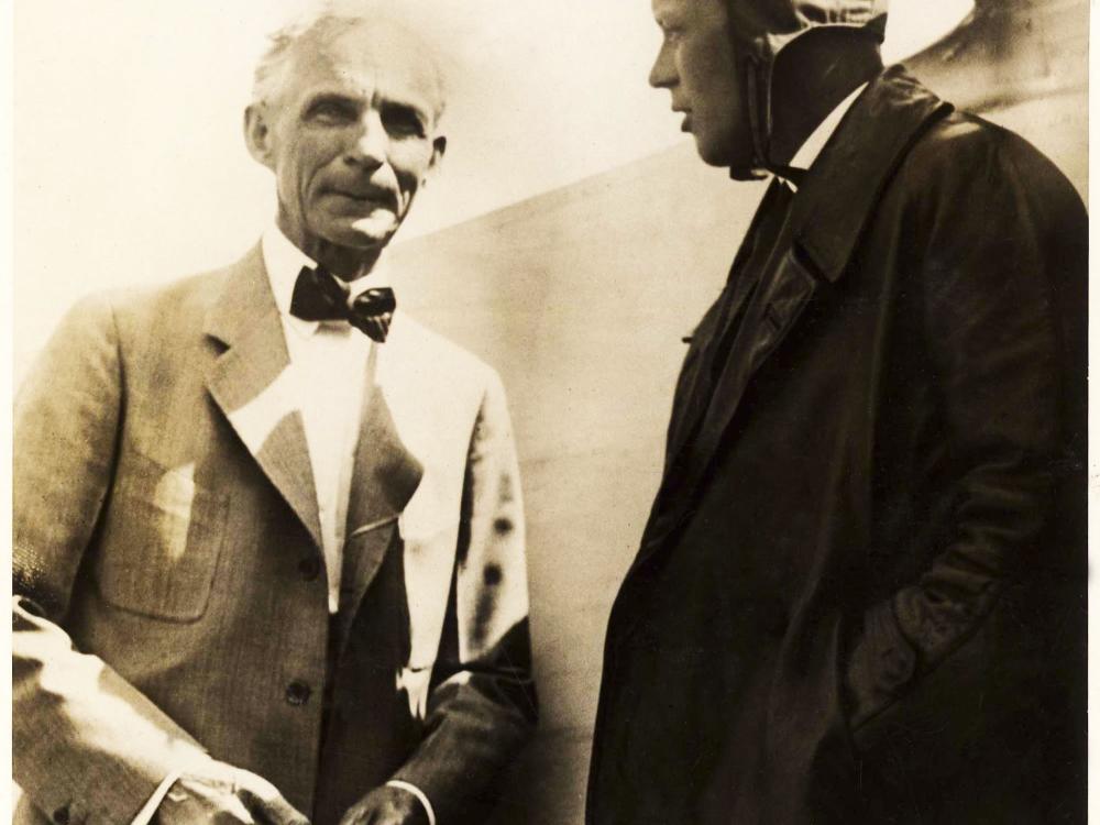 Charles Lindbergh and Henry Ford, both of whom are white men, stand together. Lindbergh is wearing aviator gear.