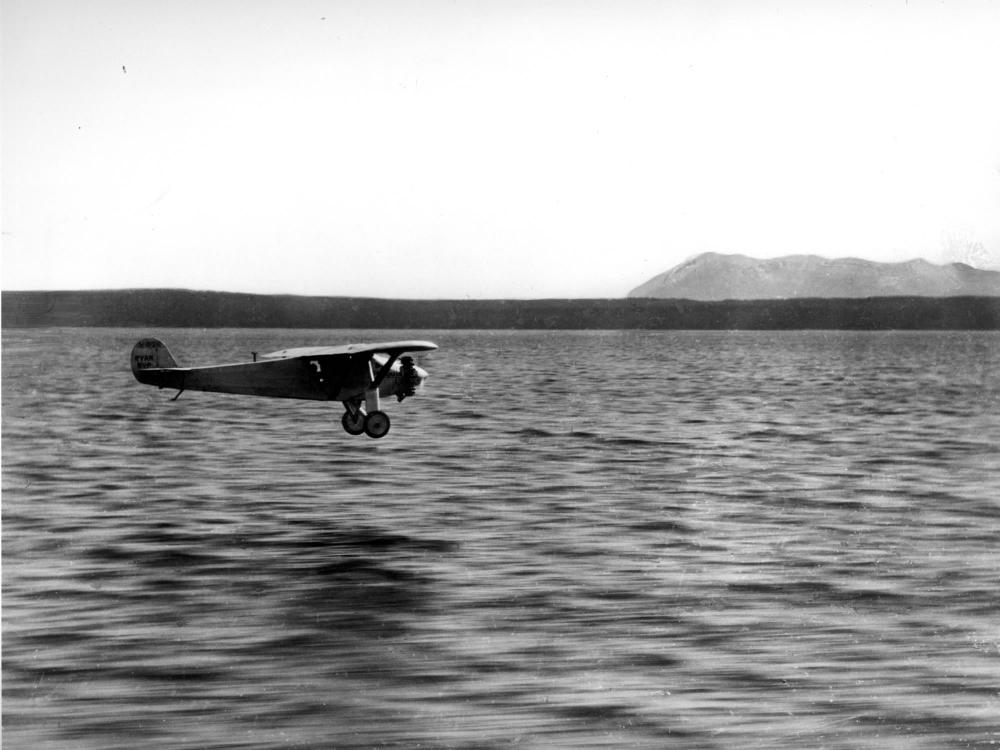 The Spirit of St. Louis, a monoplane with one engine flown by Charles Lindbergh, flies over a body of water.