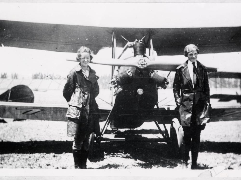 Neta Snook, a female flight instructor, stands with her at-the-time female pilot student, Amelia Earhart, in front of a biplane with one engine.