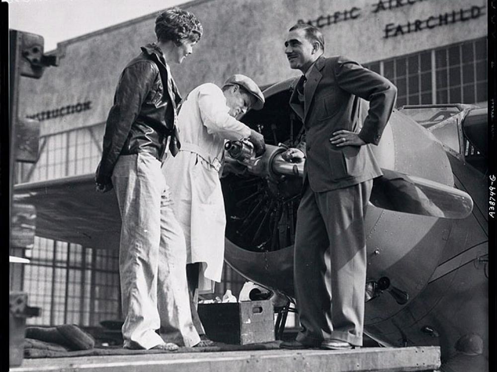 Paul Mantz, a man, stands to the right of Amelia Earhart, a white woman, as they speak to each other.