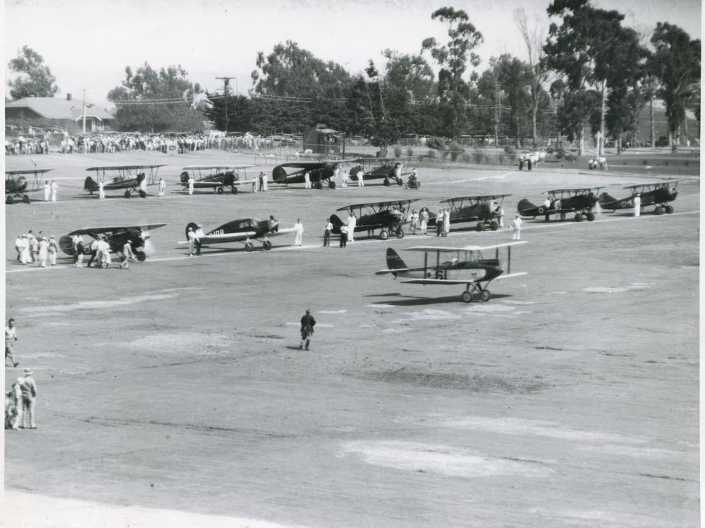 Two lines of early aircraft, mostly biplanes, are seen as part of an air race featuring only women pilots.