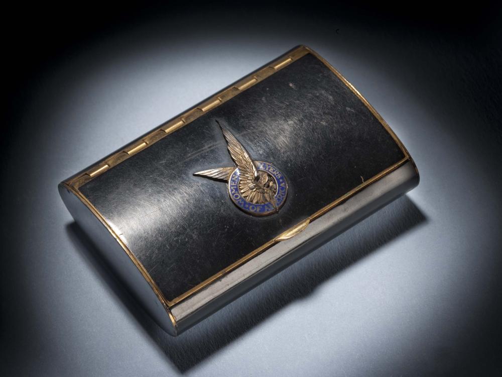 Black makeup case with closed lid. On the center of the lid, there is a silver insignia of a pair of two wings inside a blue seal.