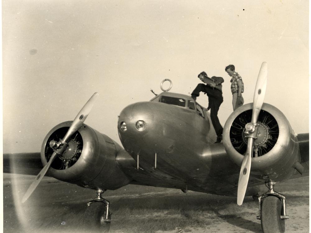 Amelia Earhart and Fred Noonan, a white woman and white male respectively, stand on top of a monoplane with twin engines. Noonan is climbing into the monoplane.