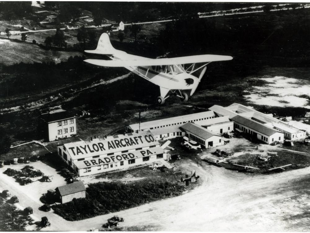 A monoplane flies over a set of buildings which are labeled as belonging to the "Taylor Aircraft Co." on the roof of a building on the left of the other buildings. Below the roof stating the company's name is the location of "Bradford, PA" on a lower roof.