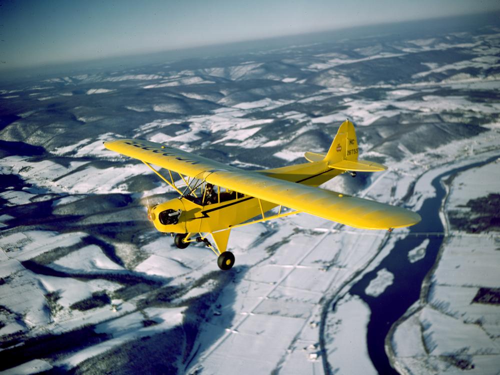 A yellow monoplane with one engine flies above land.