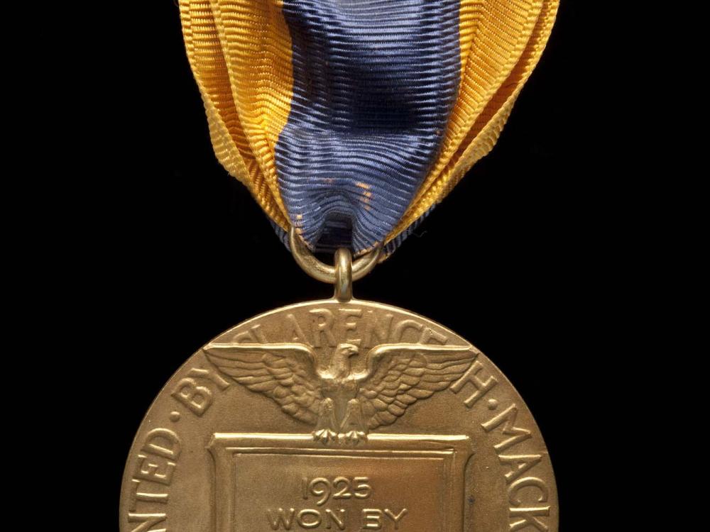 Back of aviation medal. Medallion features an etching of an eagle holding a plaque stating the winner of the medal and the rounded edges state the presenter of the medal. Suspended using yellow and black fabric attached to metal pin.