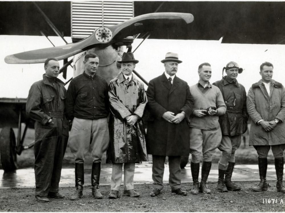 U.S. President Calvin Coolidge (a white man wearing a raincoat) stands with other white people at a meeting with pilots.